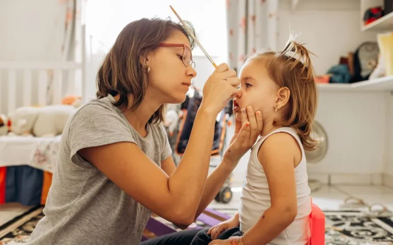 A mother meticulously face-paints her young daughter in a cozy playroom.