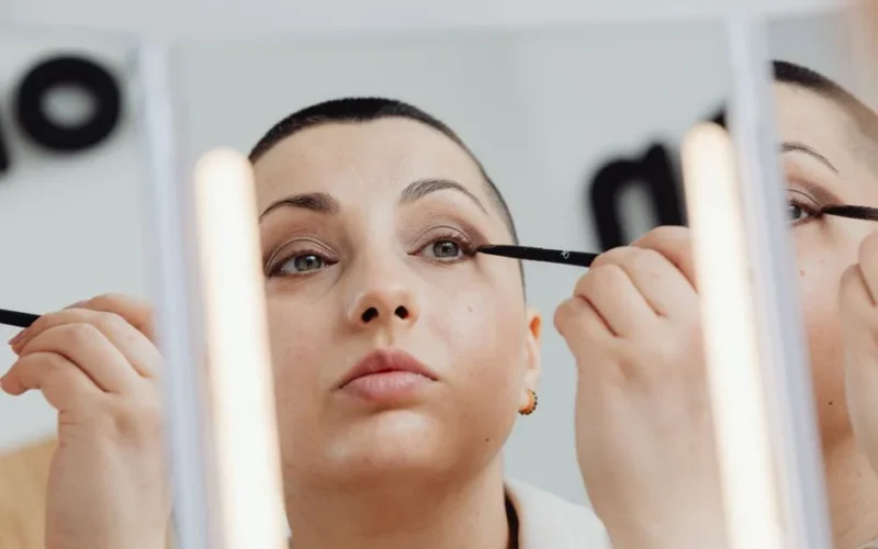 Caucasian woman with buzzcut applying eyeliner, reflected in mirror.