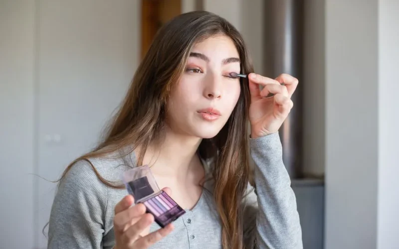 Asian woman applying makeup with eyeshadow palette indoors, showcasing beauty routine.