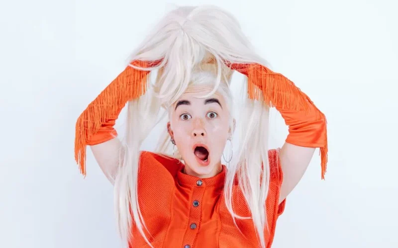 A startled woman in a vibrant orange outfit holding a blonde wig with a surprised expression.