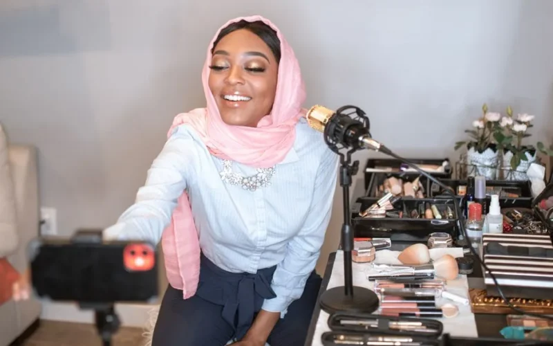 A beauty vlogger in a hijab recording a makeup tutorial with cosmetics setup.