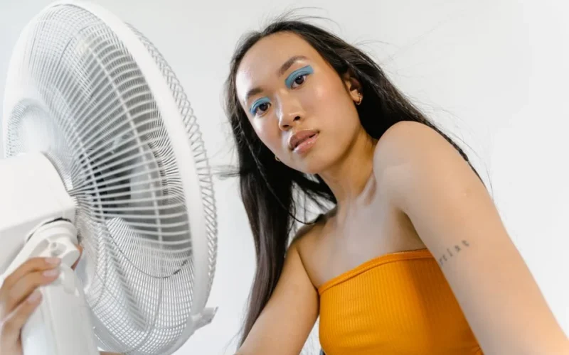 Young woman with bold makeup and tattoo adjusting a fan indoors.