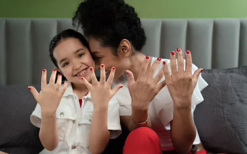 A joyful moment of a mother and daughter showing off their matching red nails while sitting together.