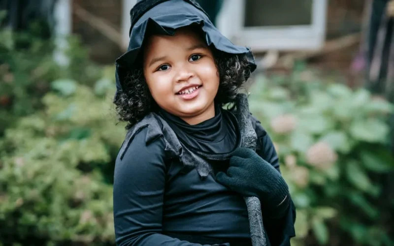 Cute Hispanic girl in witch costume and hat cuddling broomstick while smiling and looking at camera