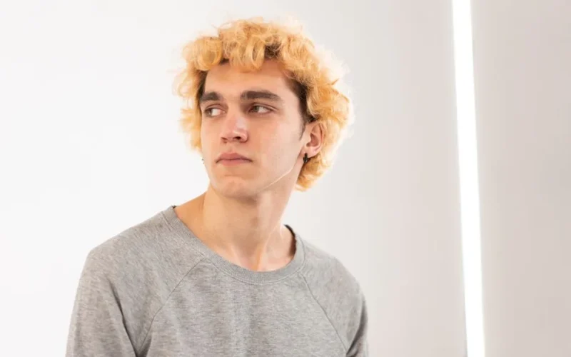 Blonde curly-haired man looks sideways in a bright indoor setting.
