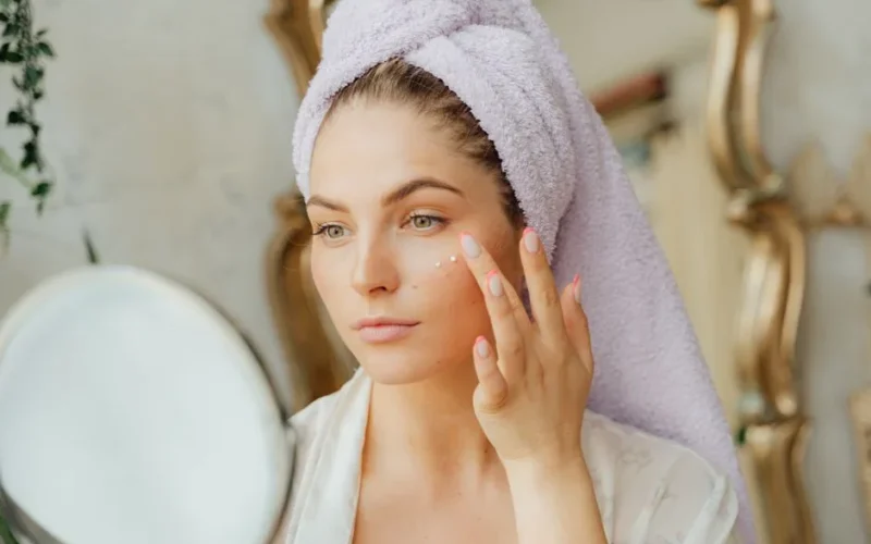 Woman with head towel applying moisturizer in front of mirror indoors.