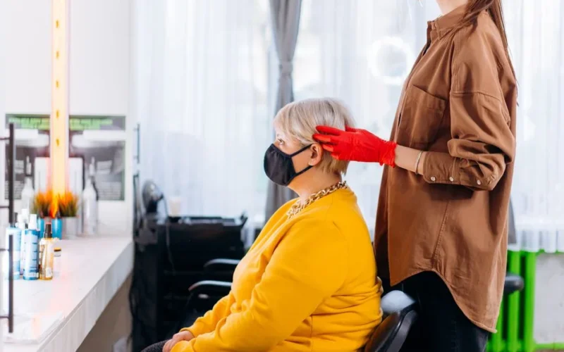 Side view of female client and hairstylist in masks and casual clothes with gloves near table with professional equipment and mirror in bright salon