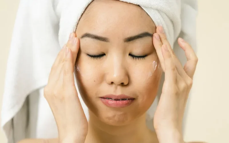 Asian woman with a towel on head applying moisturizer for skincare routine.
