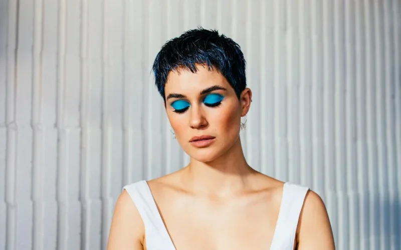 Pensive young female model with short dark hair demonstrating bright makeup with closed eyes against white wall