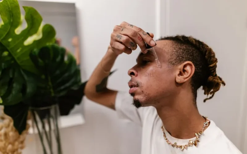 A young man with tattoos and dreadlocks applies skincare serum to his face indoors.