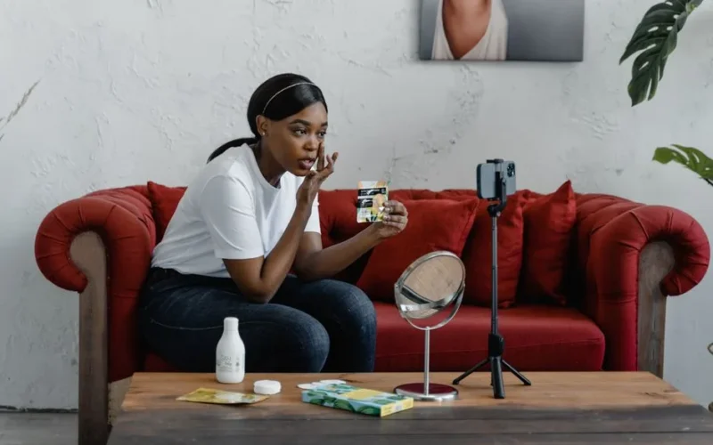 A woman records a makeup tutorial in a cozy indoor setting with cosmetics and a smartphone stand.