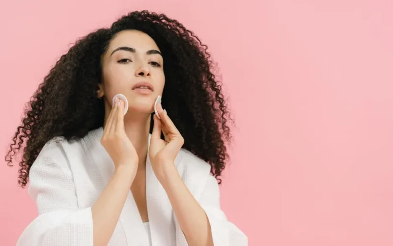 A young woman with curly hair using cotton pads for her skincare routine against a pink background.