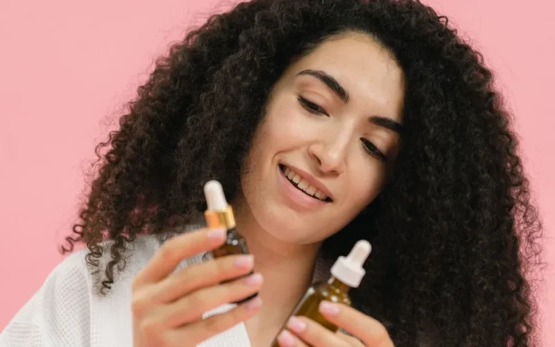 A young woman analyzes skincare bottles, set against a pink backdrop.