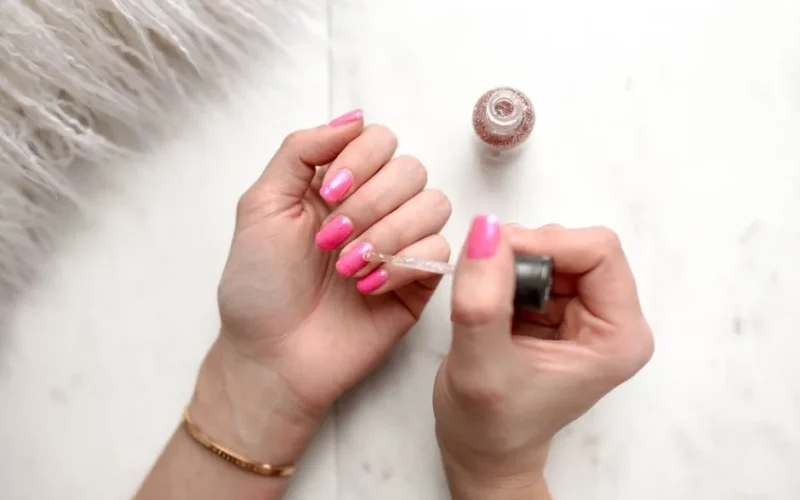 Close-up of a woman applying glitter nail polish to manicured pink fingernails.