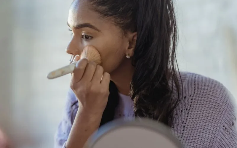 Concentrated crop African American young female spreading foundation cream on face with brush on blurred background
