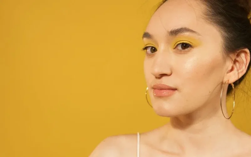 Close-up portrait of woman with striking yellow eyeshadow and hoop earrings, set against a matching yellow background.