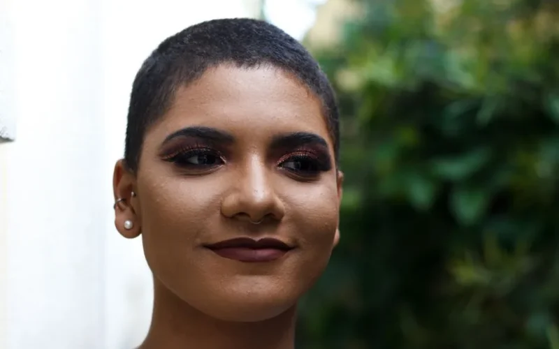 Portrait of a confident woman with bold makeup, short hair, and joyful expression in an outdoor garden setting.