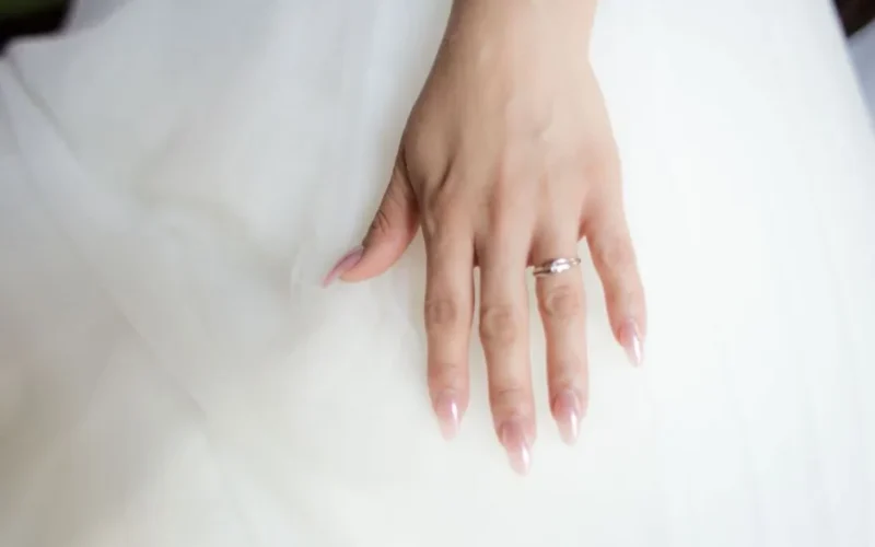 Close-up of a bride's hand with a diamond wedding ring and elegant manicure against a white dress.