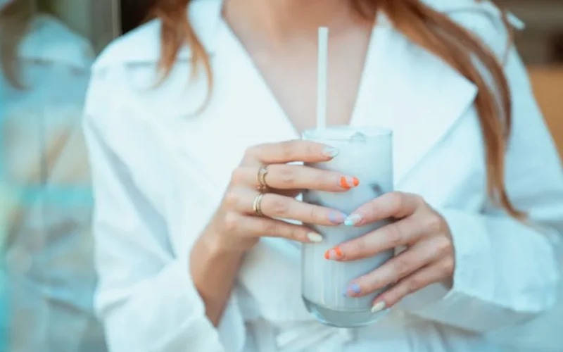 A stylish woman with colorful nails holds an iced drink in a cafe setting.