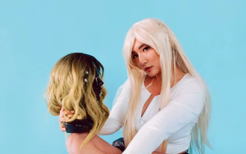 Stylish woman holding a mannequin head with a wig against a vibrant blue background.