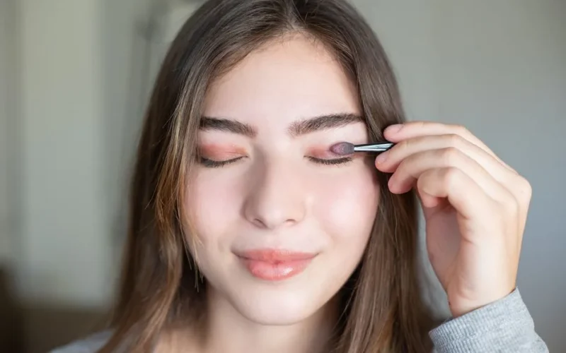 Close-up of a young woman applying eyeshadow with eyes closed indoors.