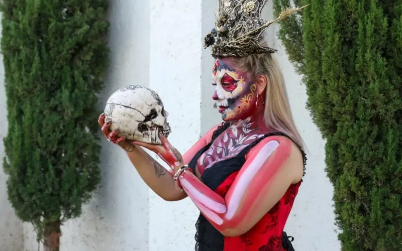 Woman in Day of the Dead makeup holding skull outdoors, vibrant costume and face paint.