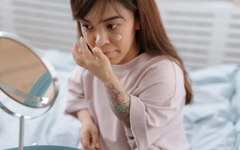 Adult woman applying face cream at home, embracing morning skincare routine in bedroom.