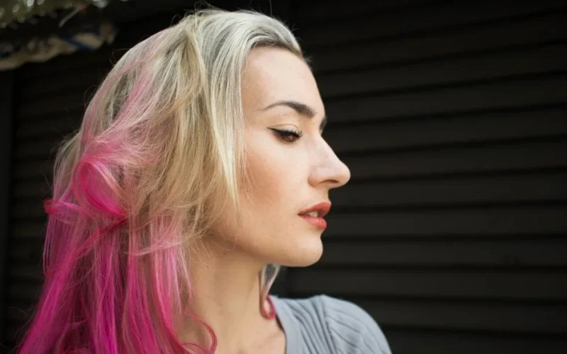Profile of a fashionable woman with blonde and pink hair in an outdoor setting.