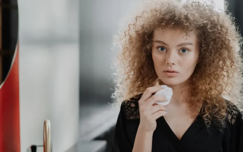 Young woman with curly hair applying beauty routine with cotton pad in a bathroom.