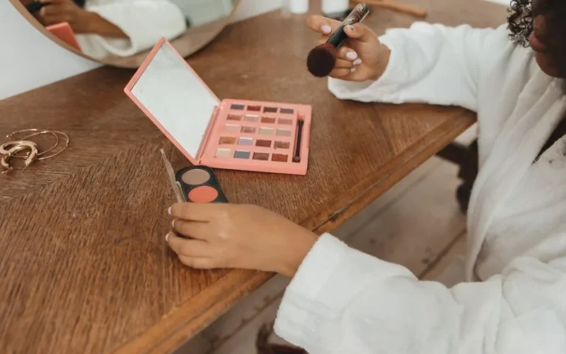 Woman wearing a bathrobe applying makeup at a home vanity table with brushes and cosmetics.