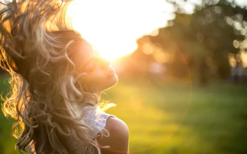 A young woman tosses her long, wavy blond hair in the sunlight during golden hour outdoors.