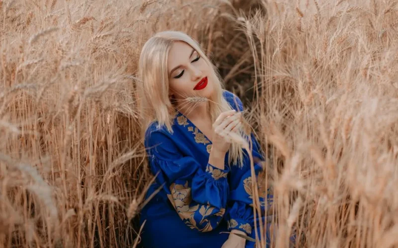 Blonde woman in a blue dress sitting in a golden wheat field during sunset.
