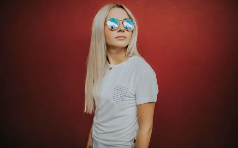 Stylish woman with blonde hair and sunglasses posing against a red background.