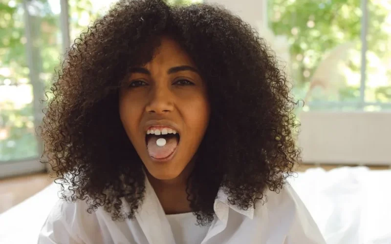 Close-up of a woman with afro hair showing a pill on her tongue indoors.