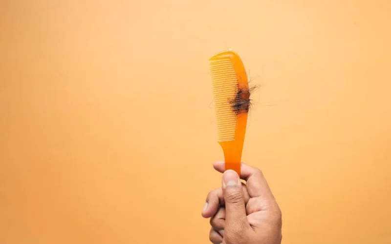 Close-up of hand holding a comb with hair against an orange backdrop, highlighting hair loss concept.