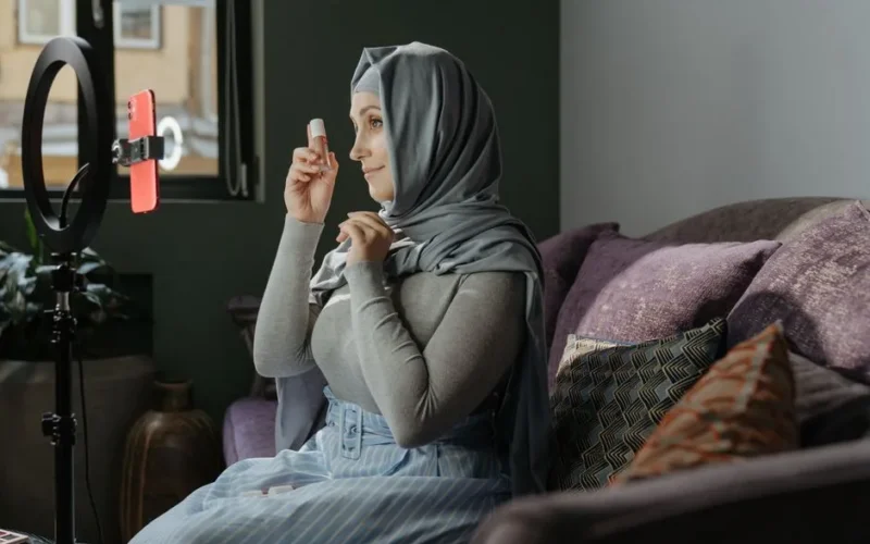 A woman in hijab applying makeup indoors, recording a tutorial with a smartphone and ring light.