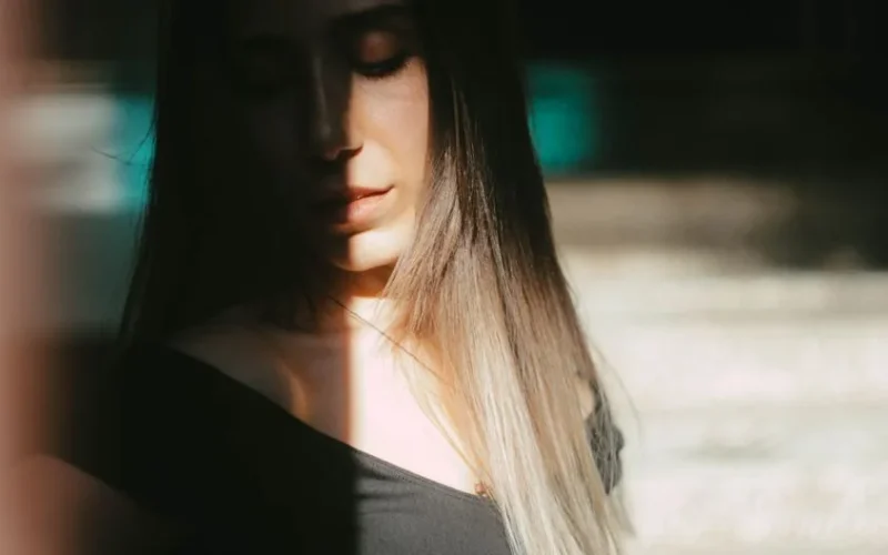 Unrecognizable female with dark hair standing with closed eyes under sunlight covering face while standing on street with blurred background