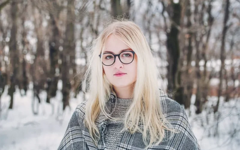 Stylish blonde woman with glasses in a snowy forest, winter fashion.