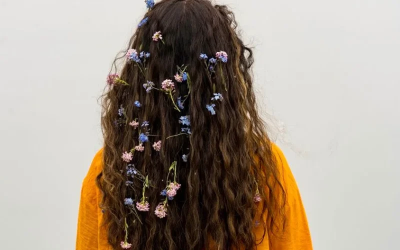 Back view of a woman with long curly hair decorated with small flowers, wearing a yellow shirt.