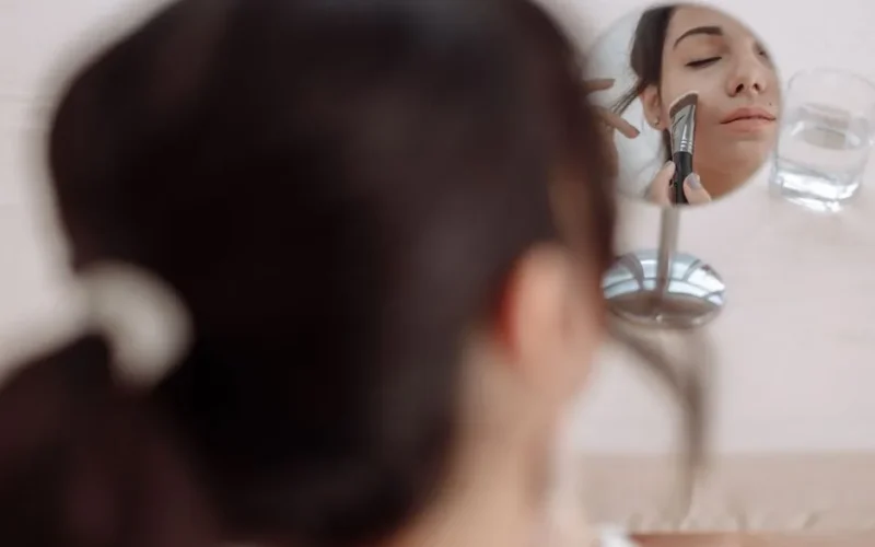 A woman applying makeup using a brush while looking into a small mirror.
