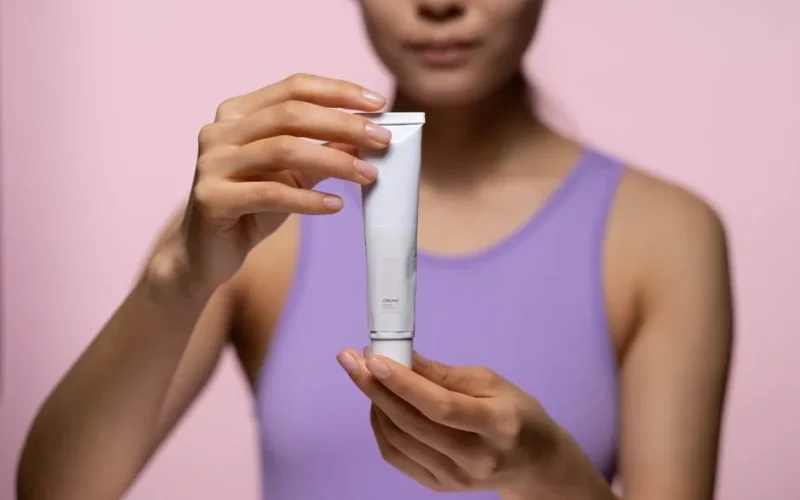 A woman in a purple tank top holds a skincare cream against a pink background in a studio setting.