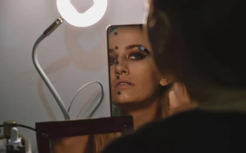 A woman applies makeup using a mirror under soft lighting, captured in a reflective portrait setting.