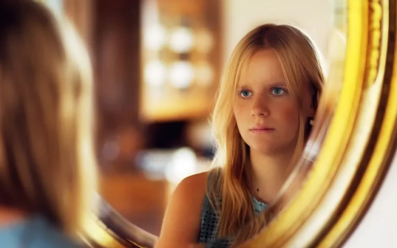 A young woman with blond hair looks at her reflection in a round mirror indoors.