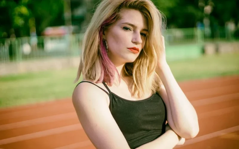 Confident young woman with blonde hair posing on an outdoor running track, enjoying the summer day.
