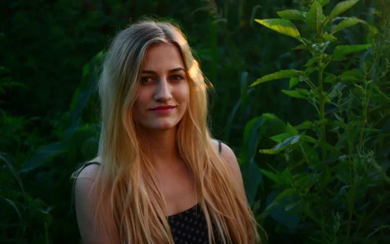 A beautiful blonde woman smiling in a natural outdoor setting with lush greenery.
