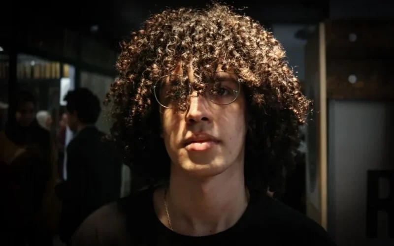 Close-up photo of a young man with curly hair and glasses, highlighting fashion style and expression.