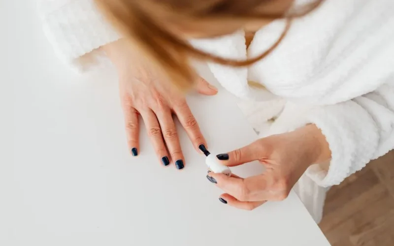 Woman in a white robe applying nail polish at home, enjoying a pampering session.