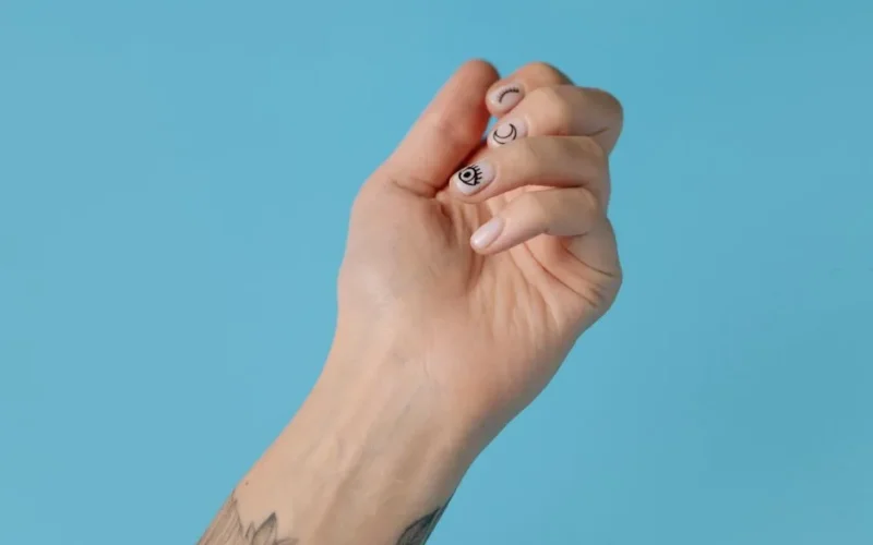 Close-up of a tattooed hand with artistic nail art against a pastel blue background, showcasing creativity and style.