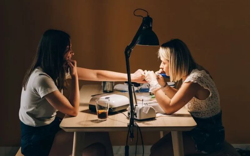 Two women at a nail salon, one receiving a manicure. Indoor setting with warm lighting.