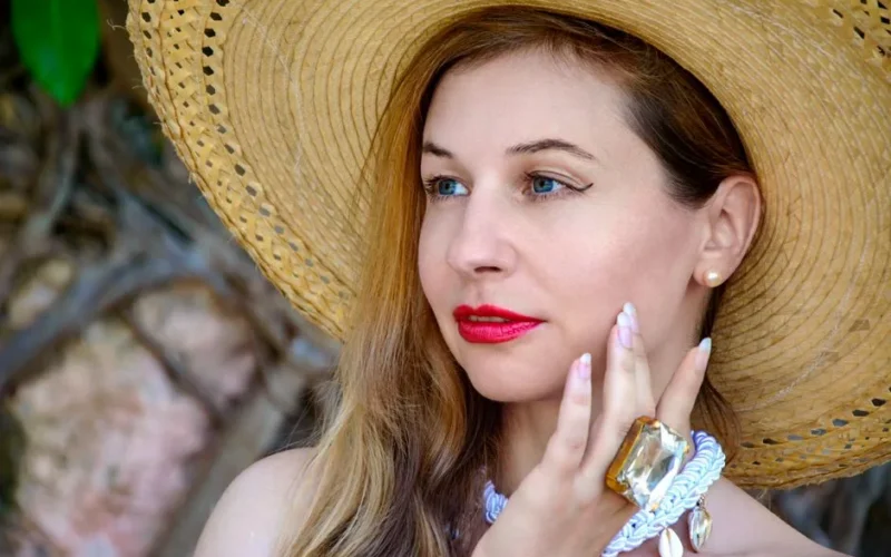 Close-up of a woman wearing a straw hat and elegant jewelry, featuring bright red lipstick and an outdoor setting.